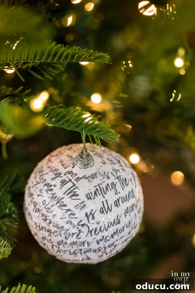El Yapımı Dekupaj Yılbaşı Süsleri 5 Grey hand writing white paper strips covering a round ball ornament on a Christmas tree. DIY decoupage ornament makeover.
