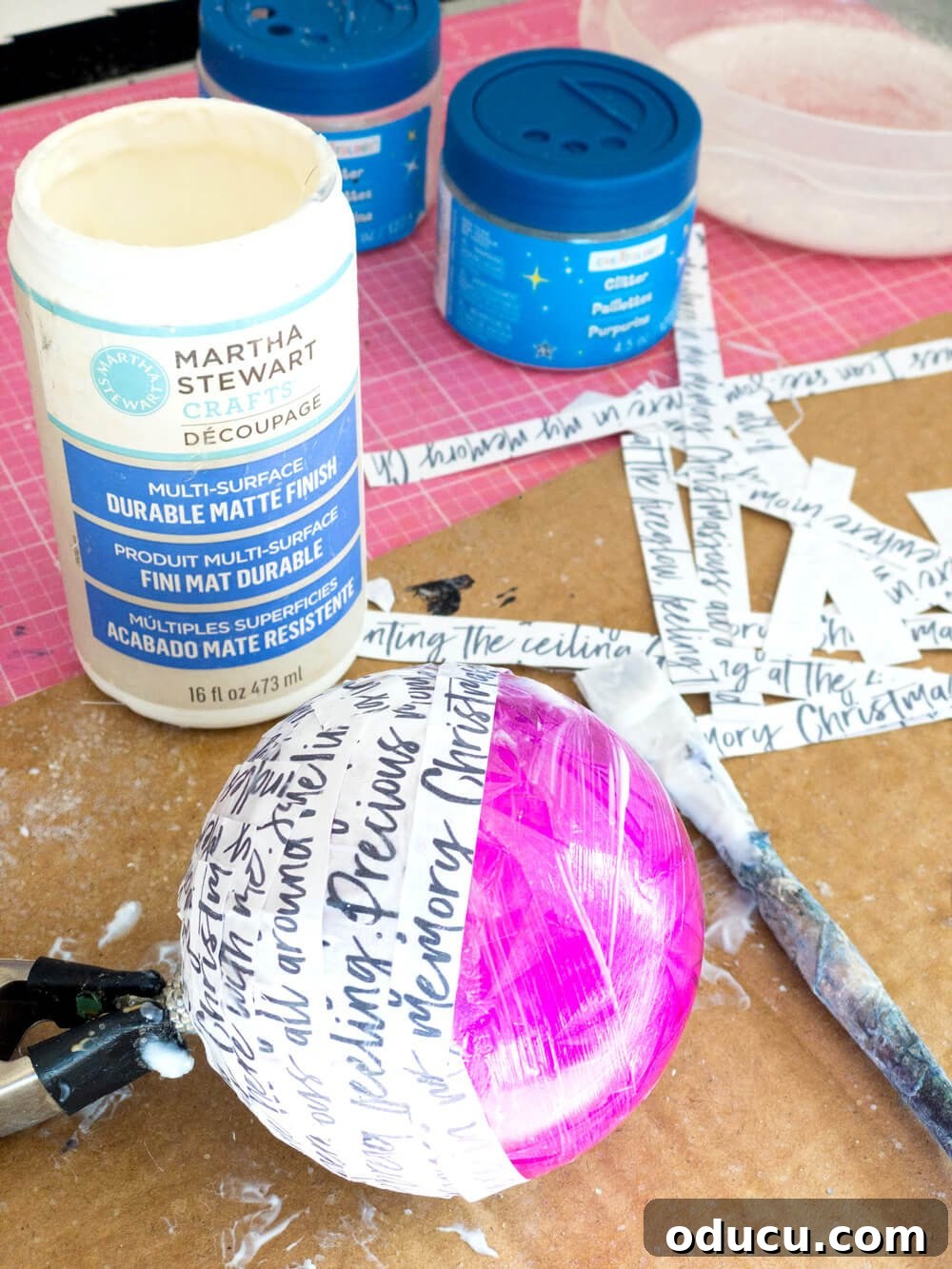 Gluing strips of hand script paper to Christmas ornaments using decoupage medium and a brush.