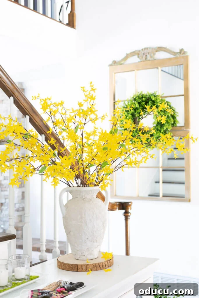 Blossom Your Entryway This Spring 4 Spring forsythia blooming in a large vase on a foyer table. A gold mirror with a green wreath hangs in the background.