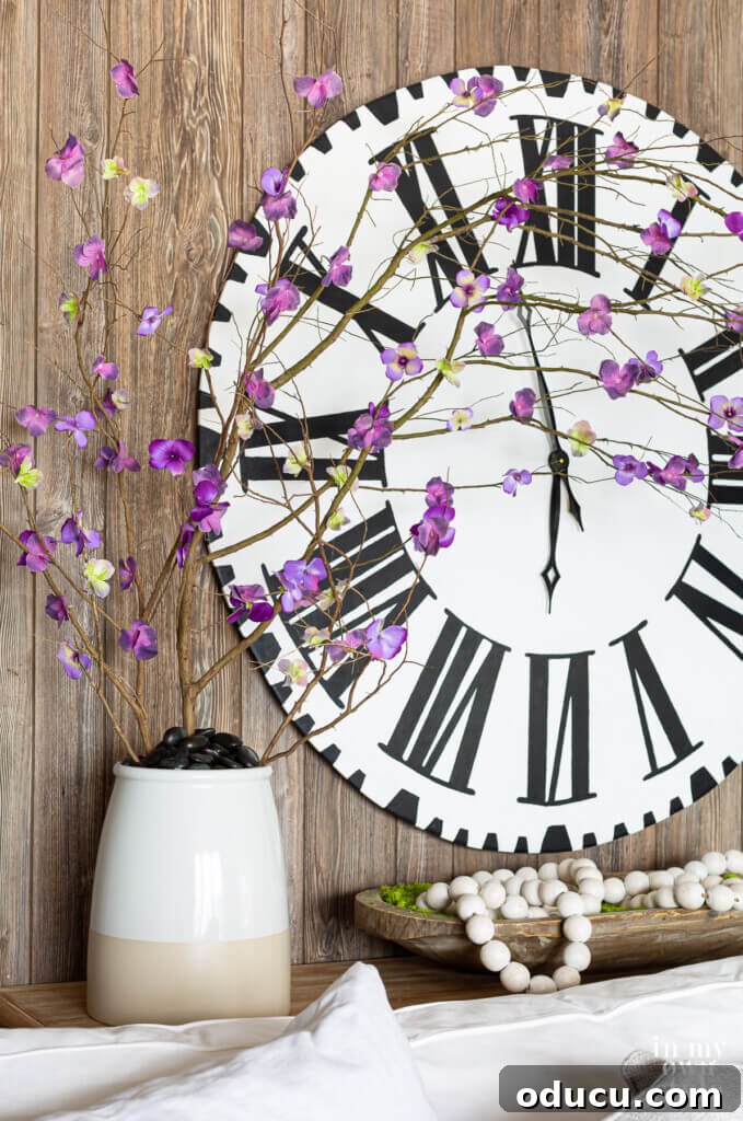 DIY Everlasting Floral Branches 3 Large Roman numeral wall clock on wall with a ceramic planter filled with faux forced branches with purple flowers.