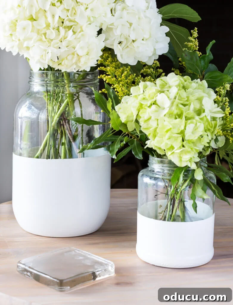 Cozy Autumn Vases: Effortless Ideas for a Warm Home 4 Two sizes of glass pickle jars transformed into modern white dipped glass vases. They are displayed on a side table.