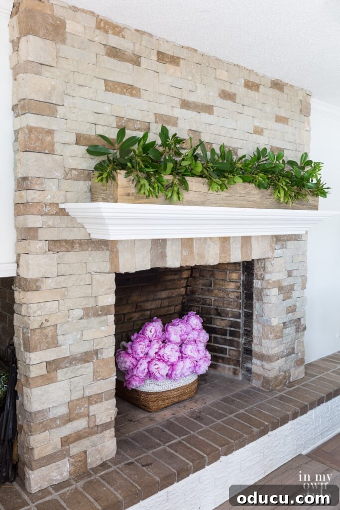Effortless DIY Fireplace Mantel 3 Stone fireplace with white mantel. Pink flowers in fireplace. Greenery clippings in wood planter on top of mantel.