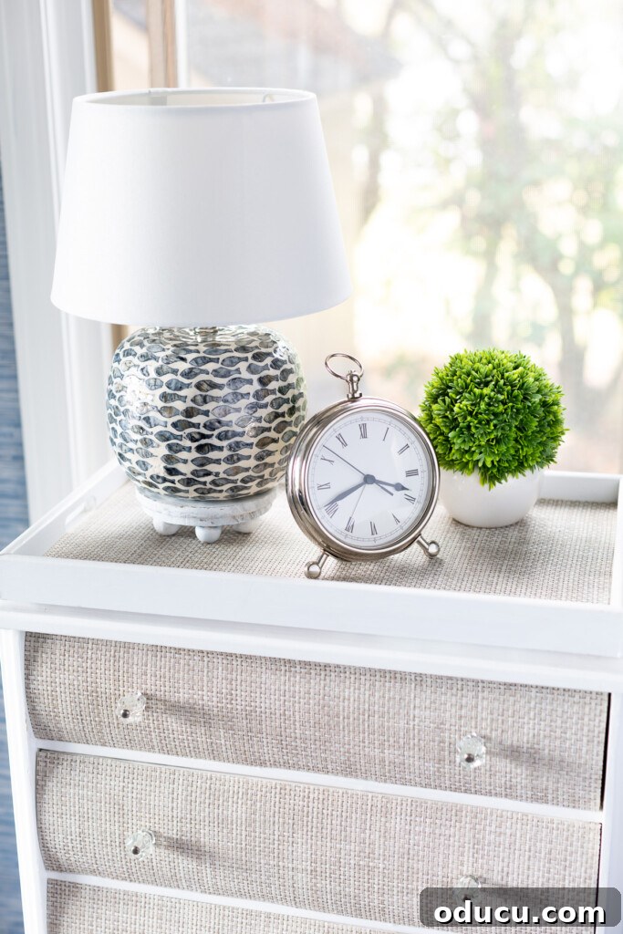 The Curious Case of My Lamp's Little Lift 11 A night table with a table lamp on a round wood riser, silver alarm clock and a small plant.