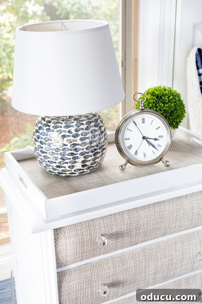 The Curious Case of My Lamp's Little Lift 4 A night table with a table lamp on a round wood riser, silver alarm clock and a small plant.