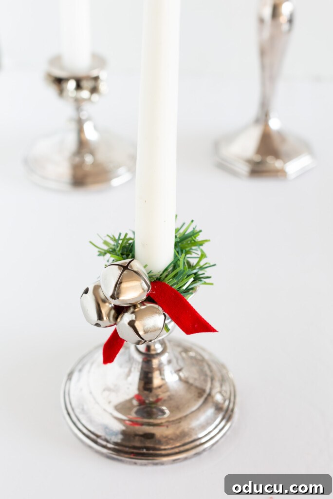 Simple Steps to Gorgeous Christmas Taper Candle Rings 12 An elegant silver candle holder with a white taper candle, featuring a DIY candle ring made from a faux greenery garland tie, a single jingle bell, and a festive red ribbon.