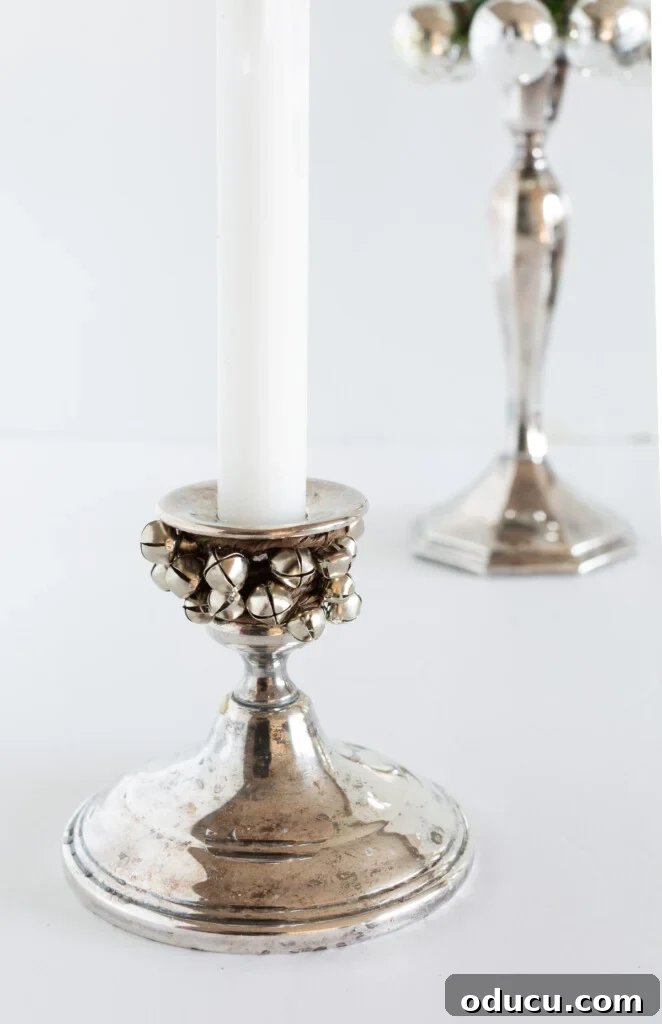 Simple Steps to Gorgeous Christmas Taper Candle Rings 13 A silver candlestick holder enhanced with a subtle garland of mini jingle bells tied around its neck, offering a whimsical touch.