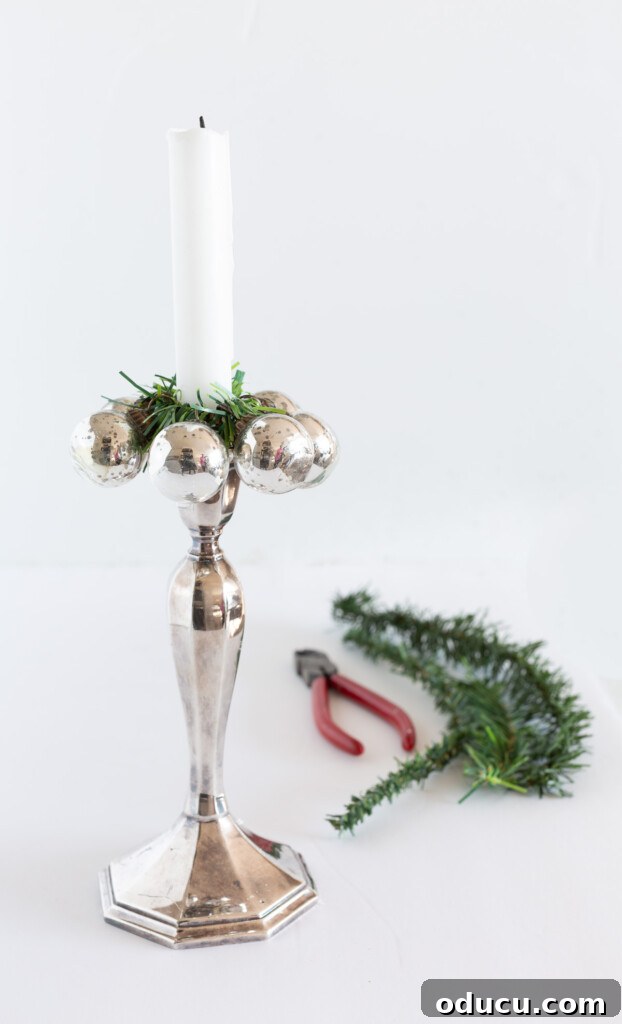 Simple Steps to Gorgeous Christmas Taper Candle Rings 6 A tall, elegant silver candlestick holder with a white taper candle, beautifully adorned with a DIY Christmas candle ring made from small silver ornaments and a garland tie.