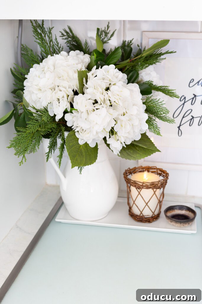 A vibrant winter white floral arrangement featuring large hydrangeas and greenery in a white pitcher, ready to brighten any home.