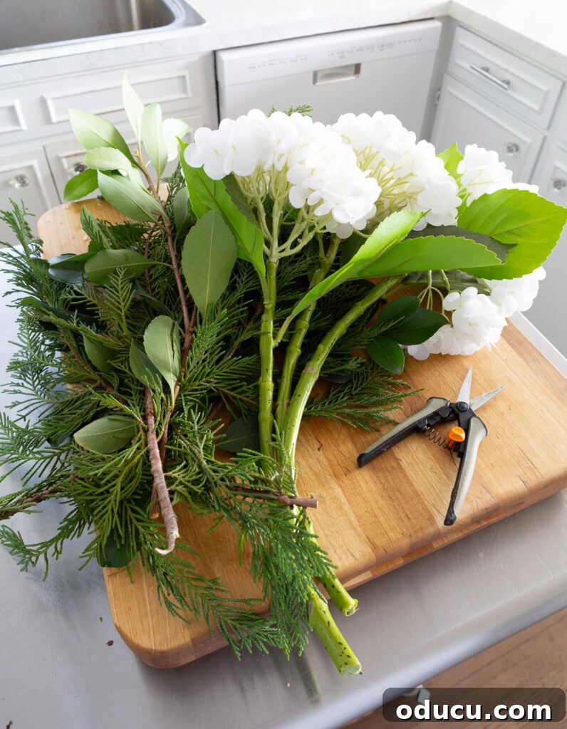 Faux white hydrangeas and evergreen clippings from my yard placed on worktable, ready for a DIY winter white floral arrangement.