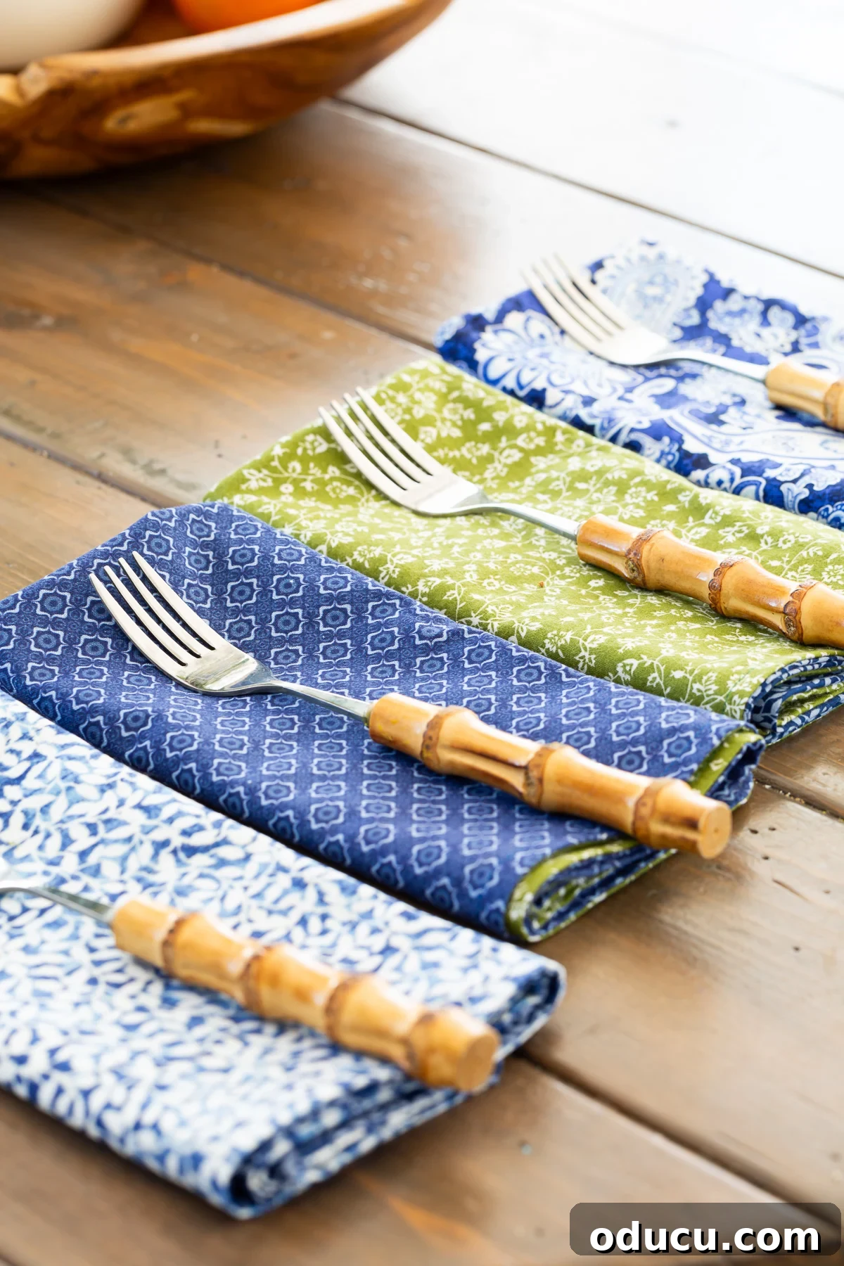 Four finished blue, green, and white reversible napkins neatly folded and lined up on a table, each with a fork, showcasing their readiness for use.
