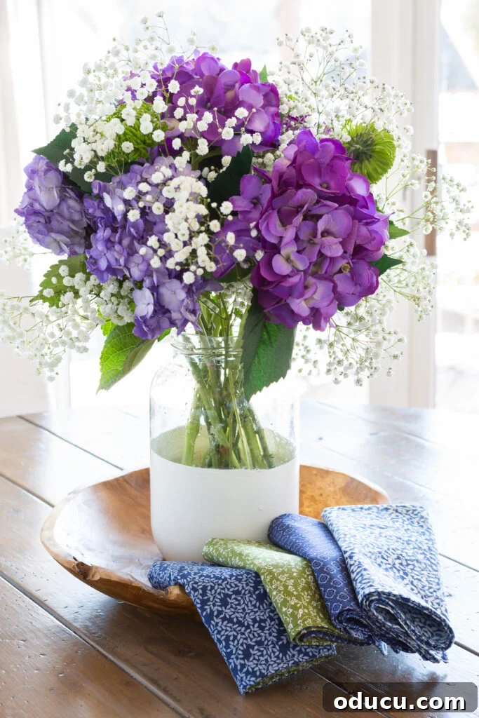 Whip Up Wonderful Napkins: A Simple Guide to Colorful Table Decor 4 A bottom-painted glass vase filled with purple hydrangeas, white, and green flowers, nestled on a wooden bowl with handmade cloth napkins folded along the edge.