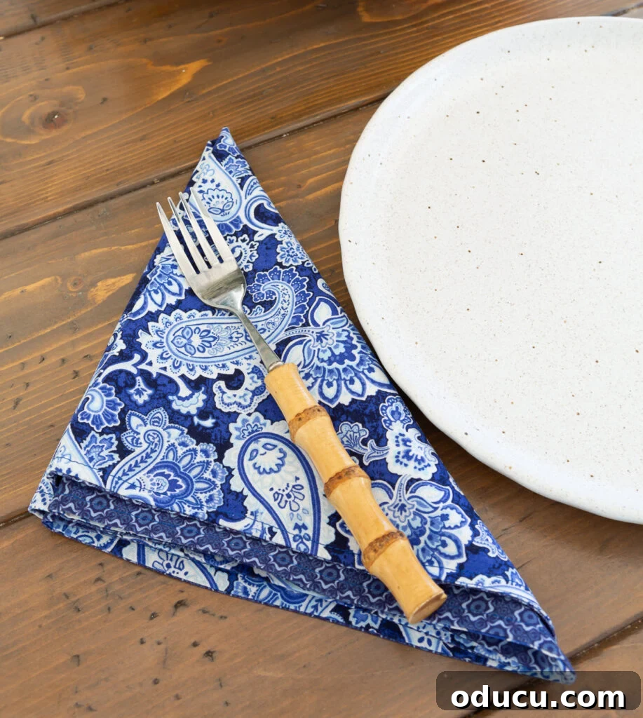 Whip Up Wonderful Napkins: A Simple Guide to Colorful Table Decor 9 A blue and white cloth napkin folded in a precise triangle shape next to a dinner plate, ready for a meal.