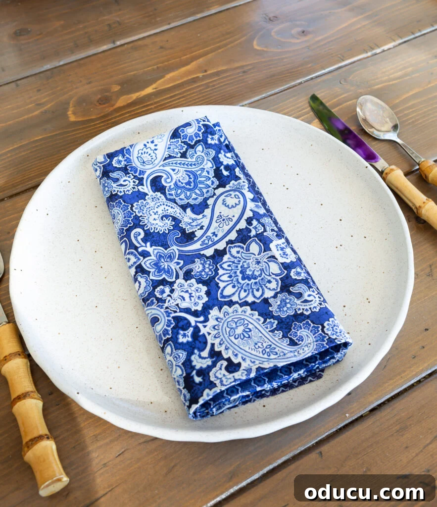 Whip Up Wonderful Napkins: A Simple Guide to Colorful Table Decor 10 A paisley blue and white cloth napkin neatly folded into a rectangle shape and placed on a dinner plate, exuding elegance.