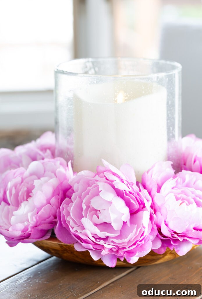 Effortless Easter Table Decor and Centerpiece Ideas 10 Close-up of a captivating Easter table centerpiece: vibrant pink faux flowers elegantly encircling a large pillar candle, all nestled within a wide, shallow bowl on a wooden table.