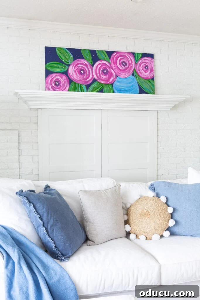 Paint Your Own Canvas: Simple Art for Everyone 3 white living room with a DIY art canvas of large pink flowers on a blue background leaning on fireplace mantel.