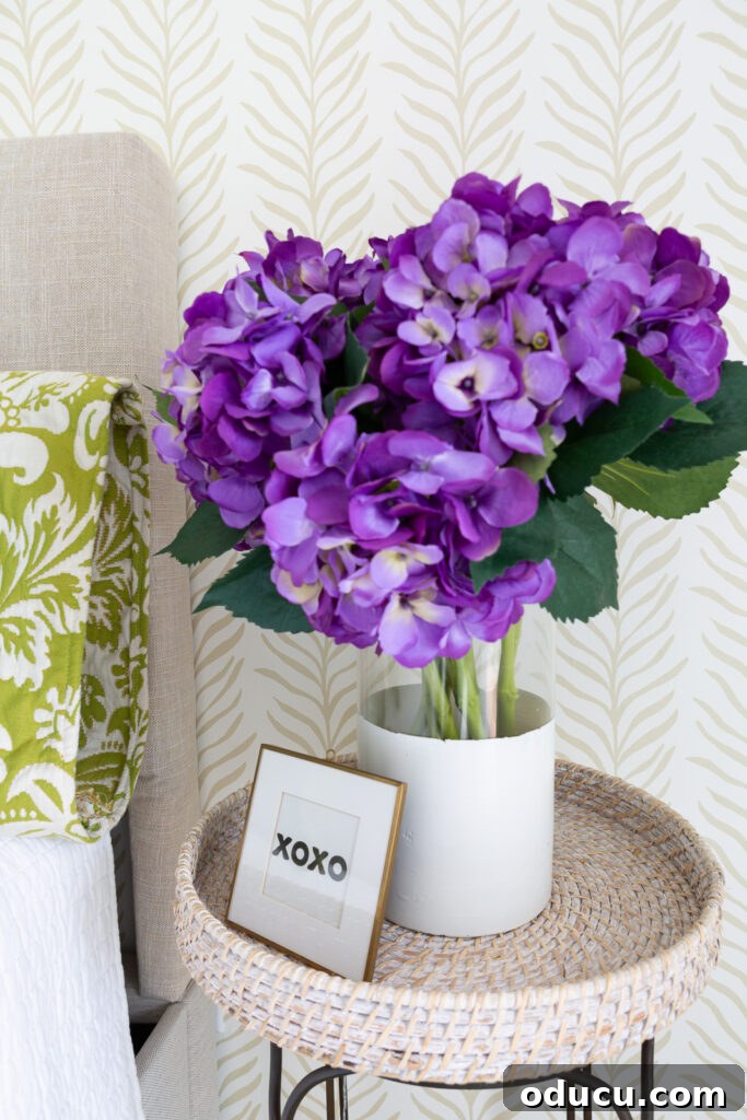 Walls of Welcome 16 A beautifully composed image featuring a glass vase holding vibrant purple hydrangeas on a table, perfectly offset by a neutral beige and white accent wall adorned with Love vs Design wallpaper.