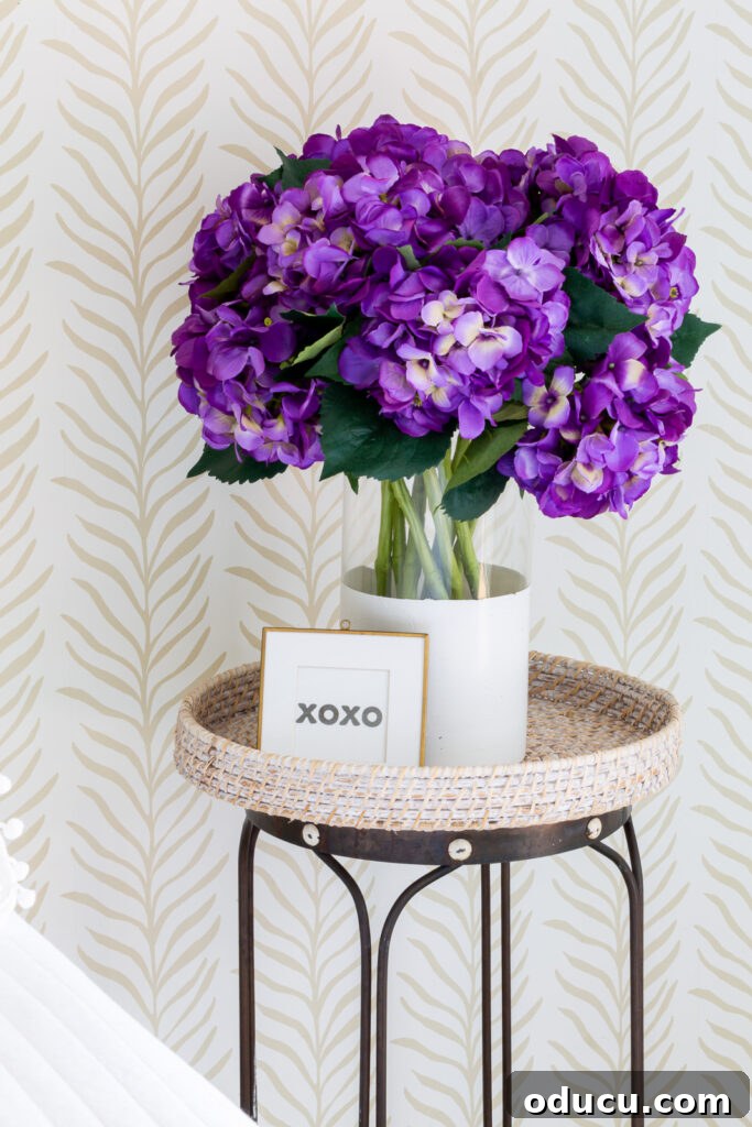 Walls of Welcome 3 A charming bedside scene featuring a vase of fresh purple hydrangeas on a side table, set against a beautifully wallpapered accent wall in a neutral Vine Thread pattern from Love vs Design, blending seamlessly with bedroom decor.