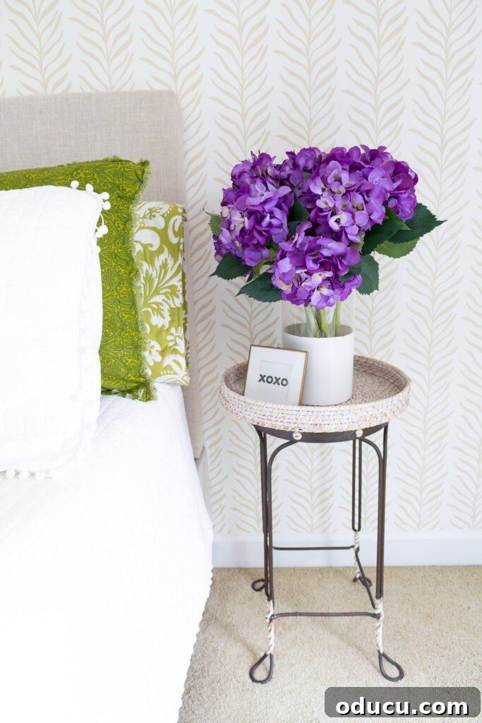 Walls of Welcome 8 A charming vignette featuring a clear glass vase overflowing with vibrant purple hydrangeas, positioned on a table against a backdrop of a neutral beige and white accent wall, adorned with Love vs Design wallpaper.