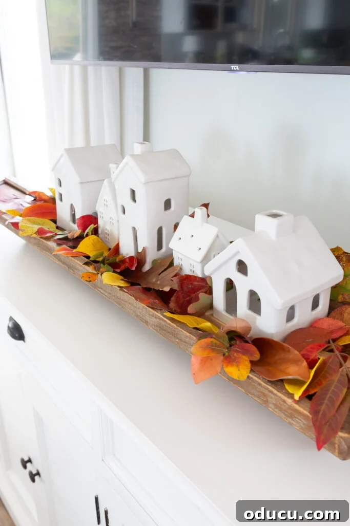 Tealight Village Magic: Fall and Holiday Decor 5 A charming ceramic Christmas village nestled among colorful fall leaves in a long dough bowl, creating beautiful autumn decor for a home.