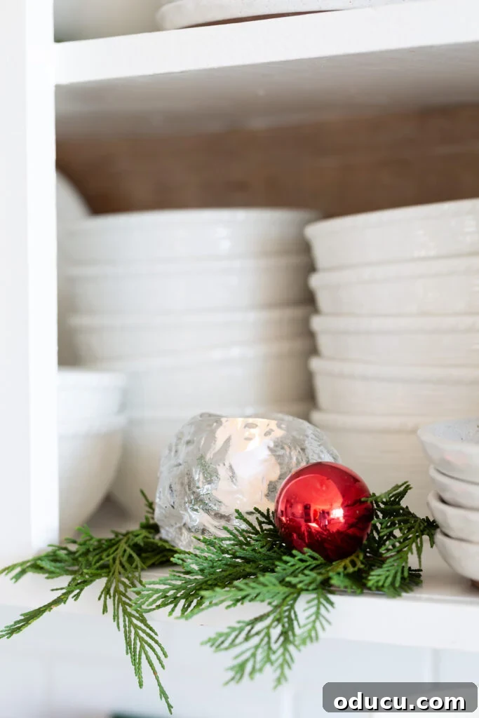 Decking the Halls for the Holidays 14 Kosta Boda snowball votive holder on an open kitchen shelf with greenery around it and a red ball ornament.