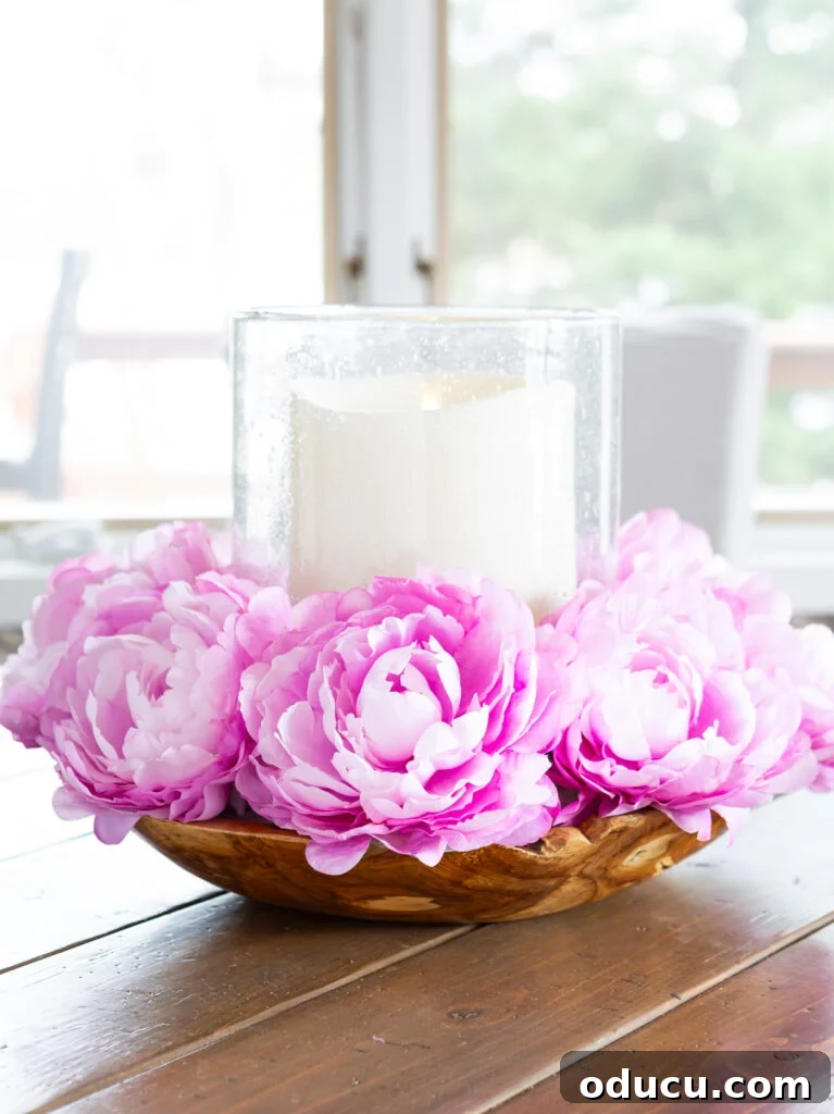 Pink flowers surrounding a large candle that is placed in a large bowl on a wood table.
